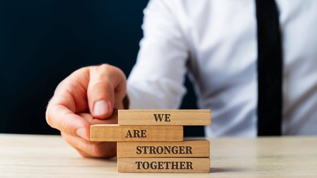 A hand placing a wooden block labeled "TOGETHER" on a stack of blocks reading "WE CAN ENHANCE SERVICES," with a person in a white shirt and black tie in the background.