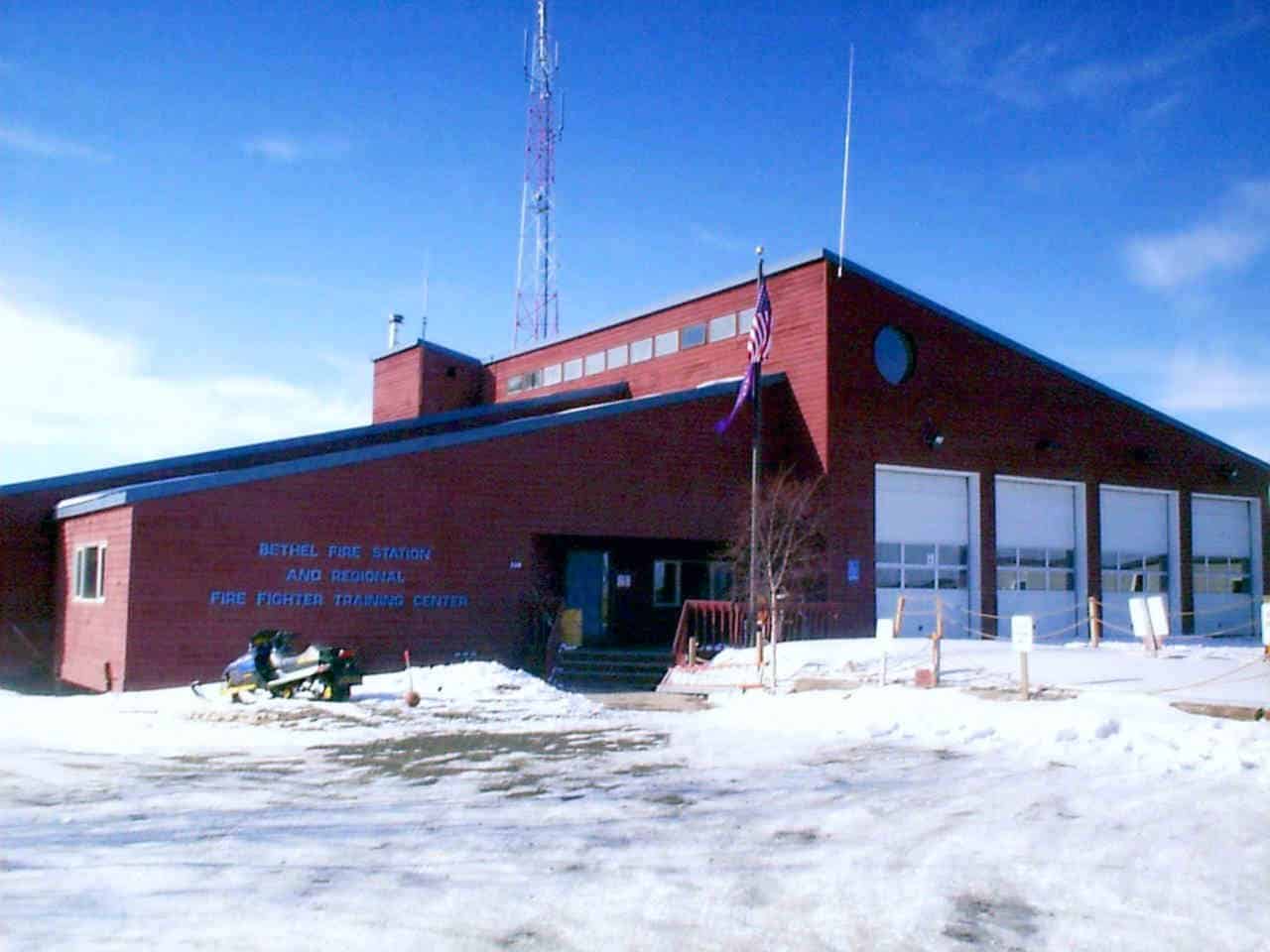 A red building with multiple garage doors, identified as the Bethel Fire Station and Regional Firefighter Training Center, surrounded by snow, with a snowmobile parked nearby and an American flag flying.