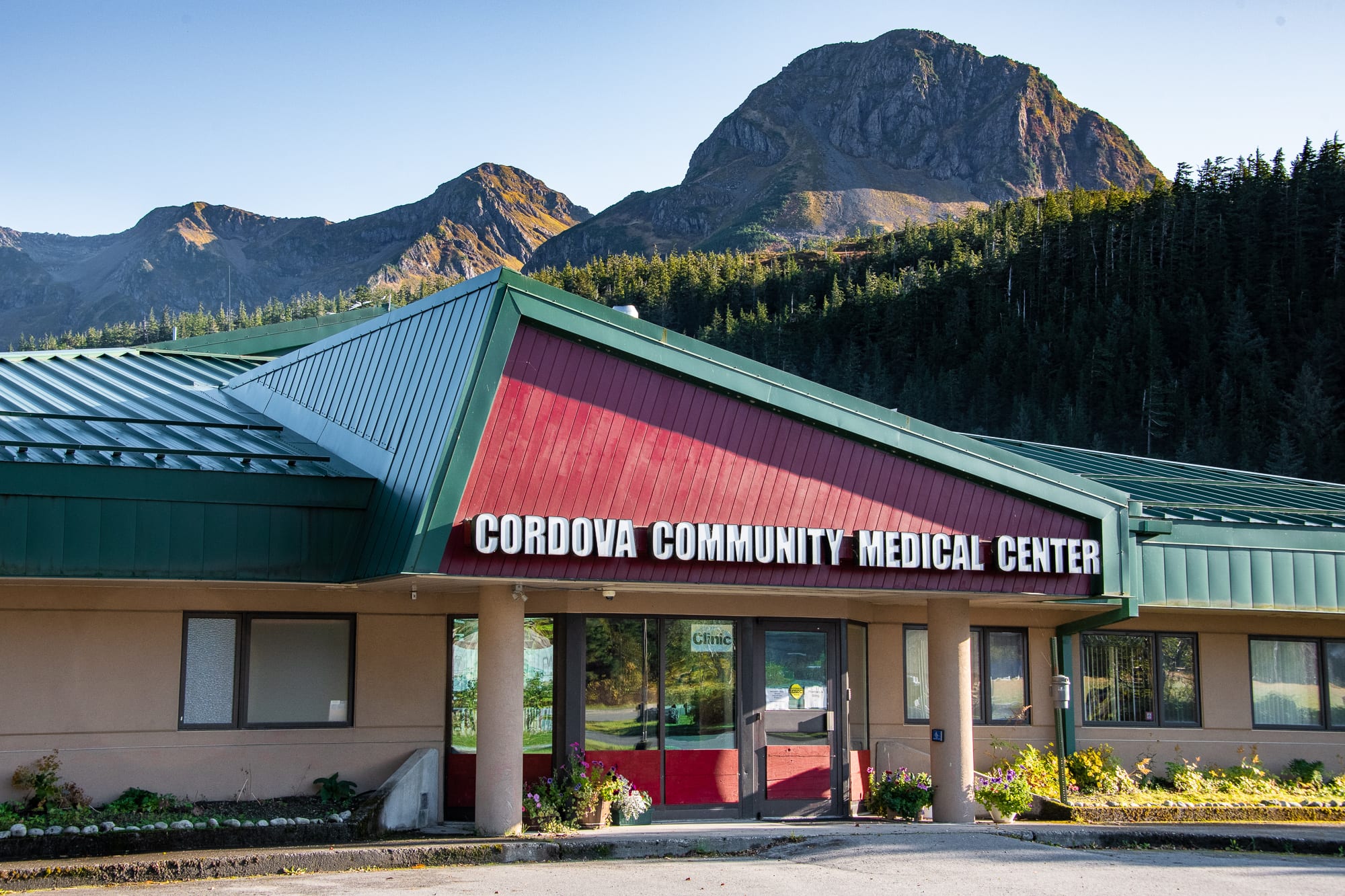A medical center building with a red and green roof, displaying "Cordova Community Medical Center" signage, set against a backdrop of forested mountains under a clear sky.