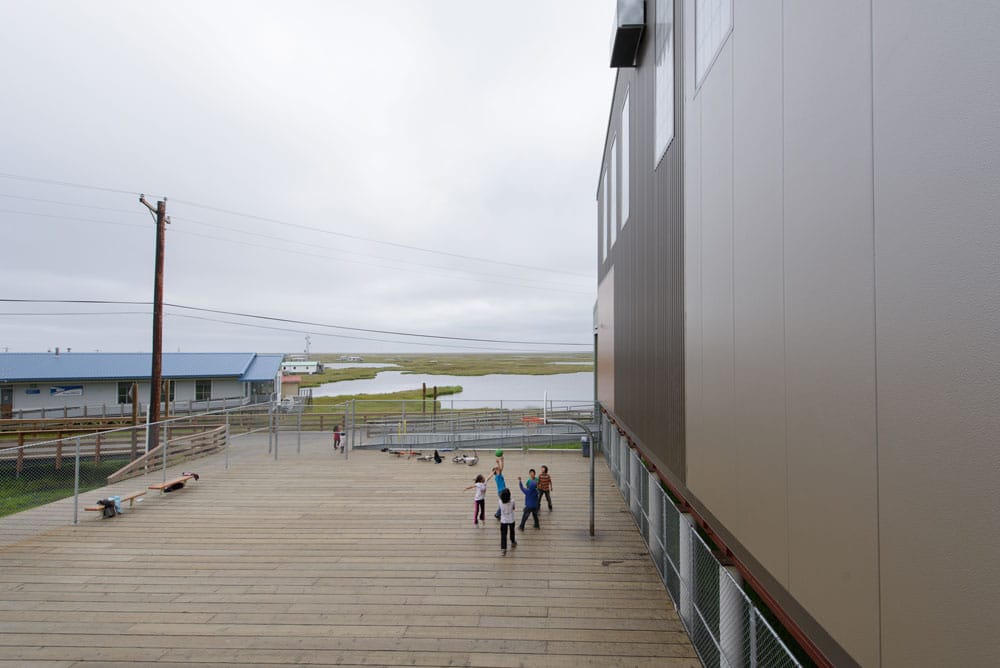 Children playing basketball on a wooden court next to a modern building with a gray exterior, overlooking a marshy landscape with water and distant buildings under a cloudy sky.