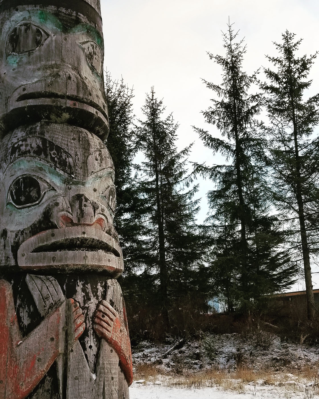 A close-up of a weathered totem pole with carved figures, set against a backdrop of tall evergreen trees and a light covering of snow on the ground.