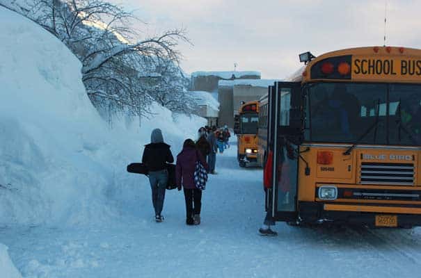 A yellow school bus with its door open, parked on a snowy path, with students walking away from it towards a building in a snowy landscape.